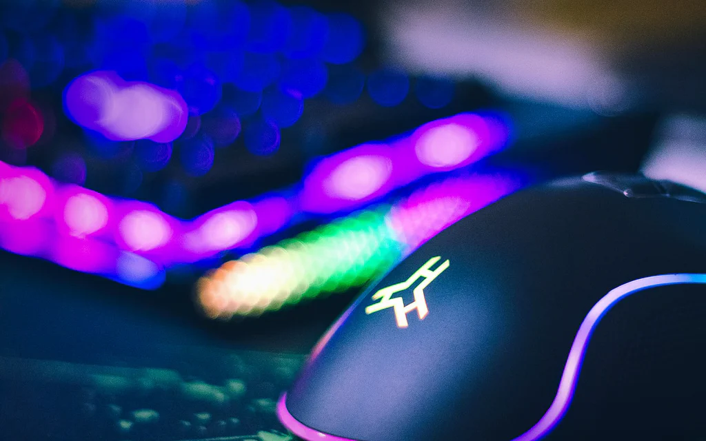 Close-up RGB gaming mouse on a dark desk with colorful keyboard lighting in the background.